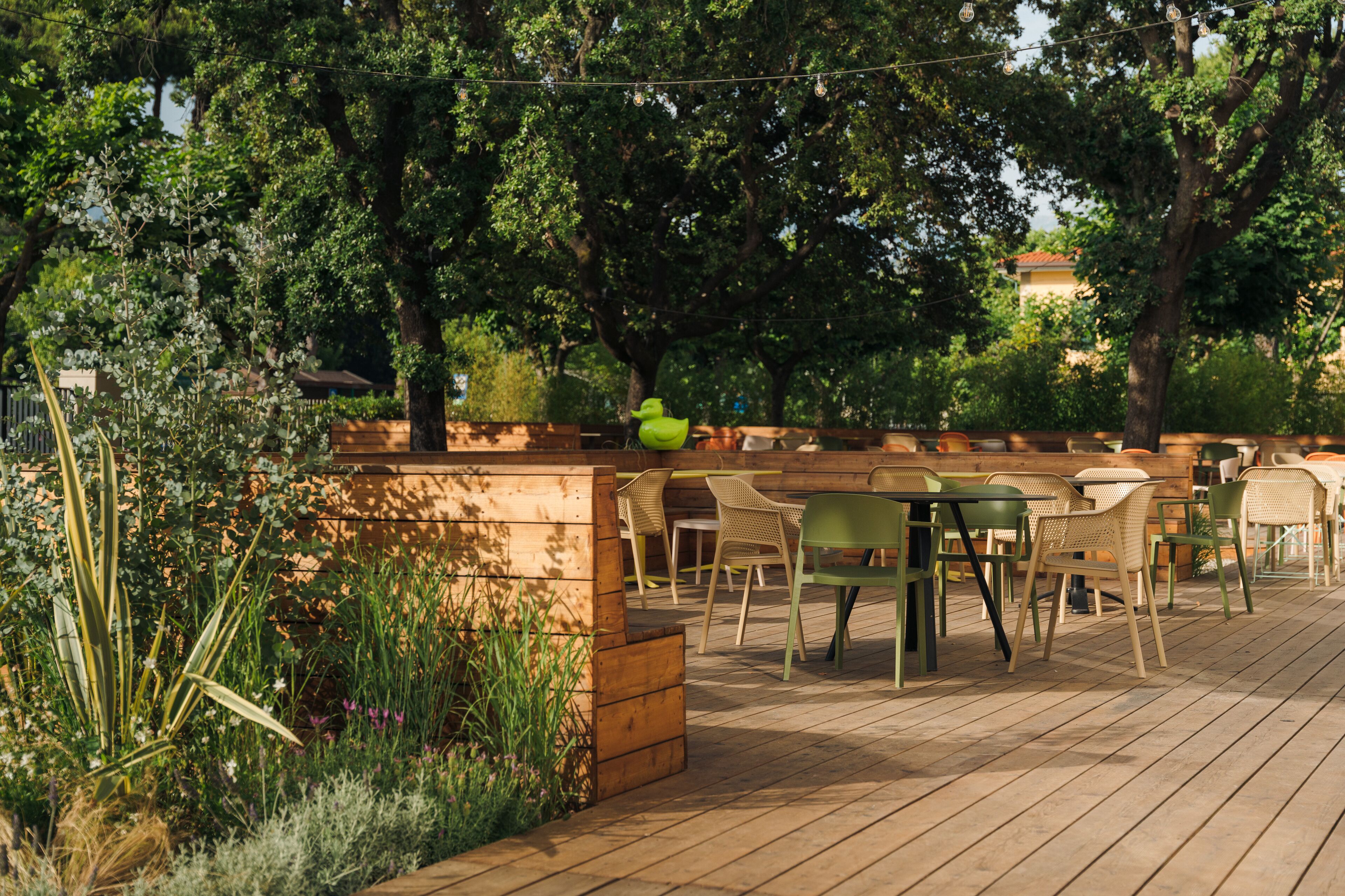 Un patio al aire libre con mesas y sillas de madera, rodeado de follaje y árboles.