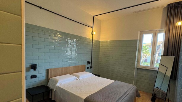 Ein Einzelbett mit Holz-Kopfteil, ein Nachttisch, ein Spiegel und ein Fenster mit Vorhängen.