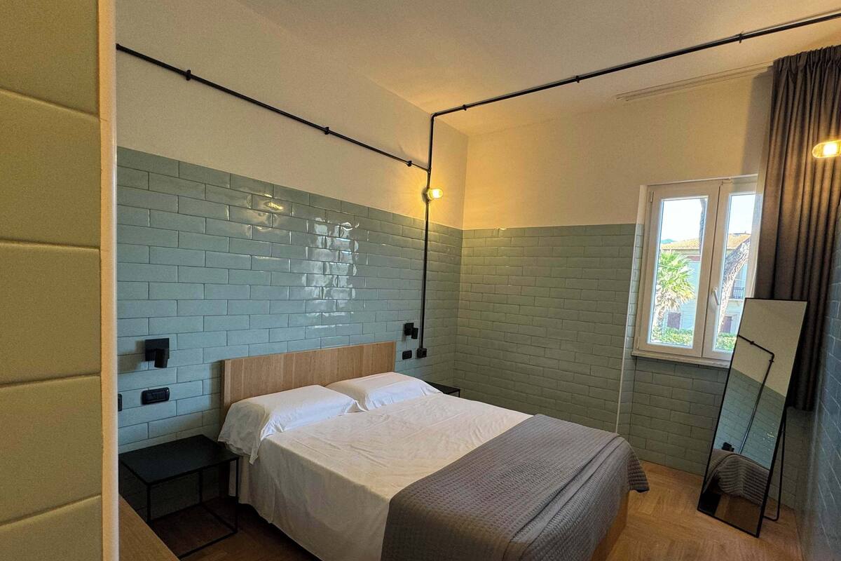Un lit simple avec une tête de lit en bois, une table de chevet, un miroir et une fenêtre avec des rideaux.
