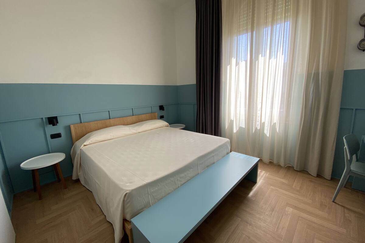 Une chambre bien rangée, avec un lit, des tables de chevet, un banc et une fenêtre avec des rideaux.
