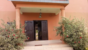 A building entrance with a dark wooden door, a small overhang, and two potted plants with red flowers. - Hôtel Lobé (Kribi)