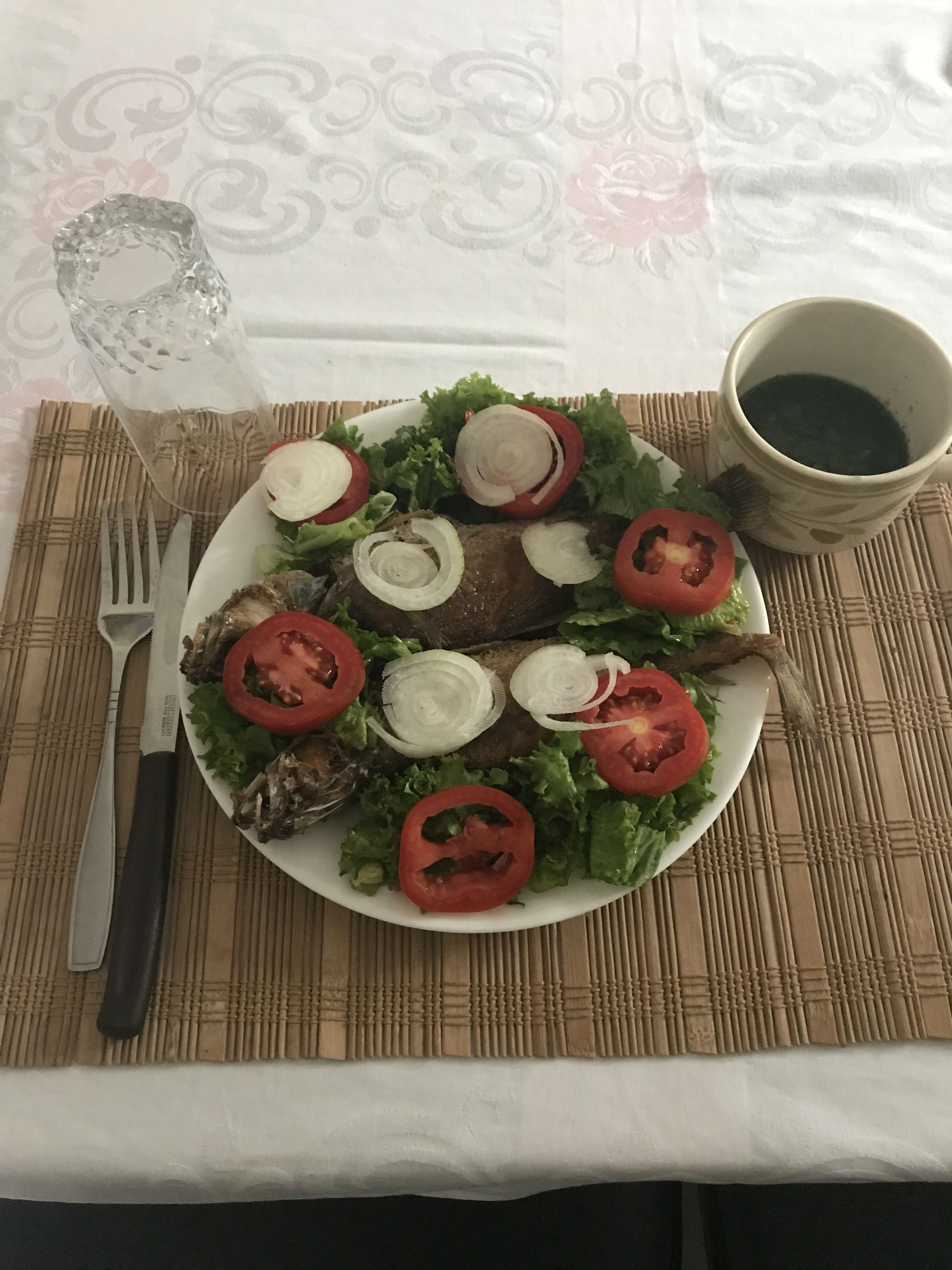 Ein Teller mit Fisch, Salat, Tomaten, einer Gabel, einem Messer und einer Tasse Tee auf einem Tisch mit gemustertem Tischläufer.
