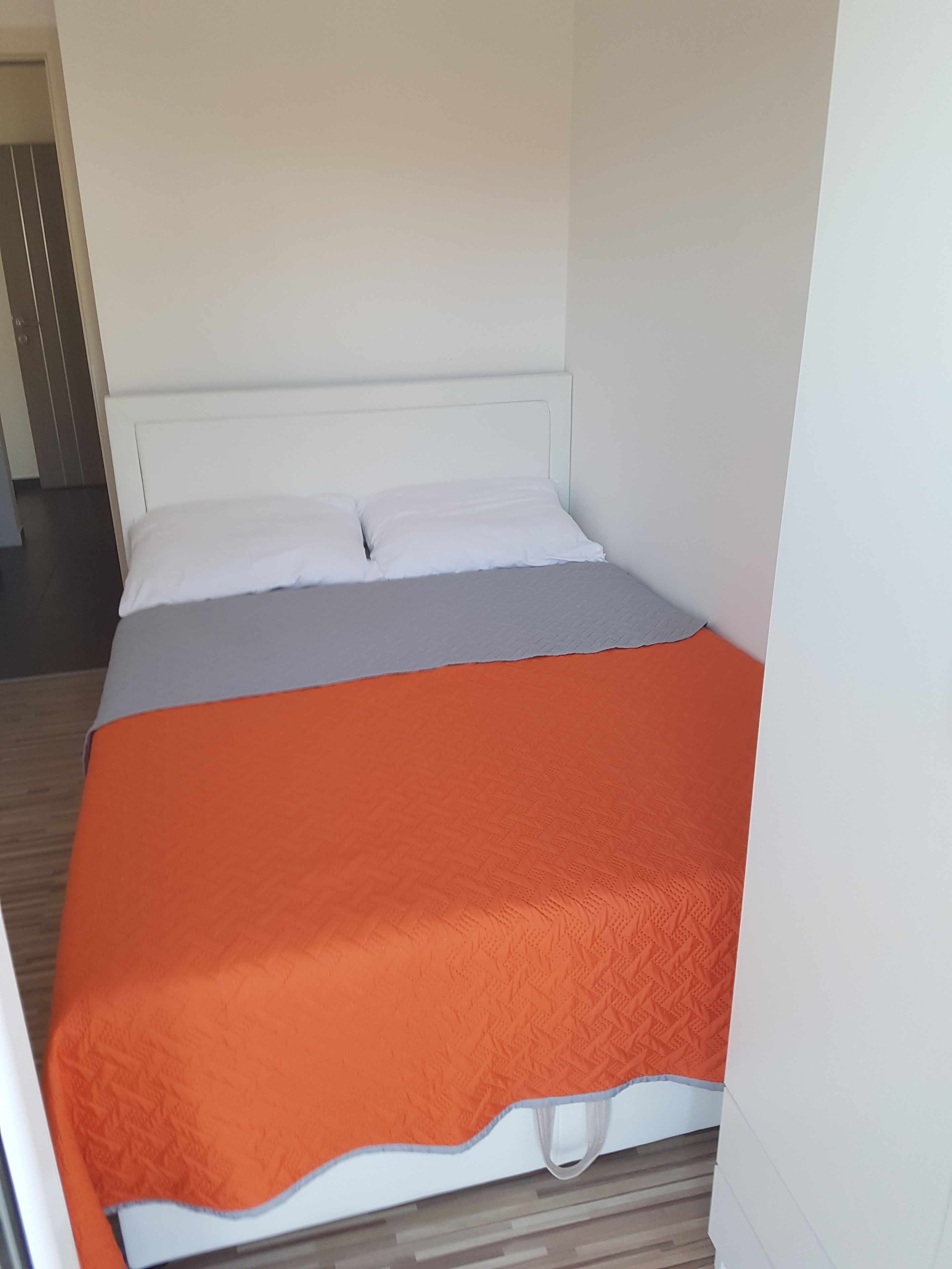 Un lit simple avec une literie blanche et orange, dans une pièce dotée d’un plancher en bois et de murs blancs.