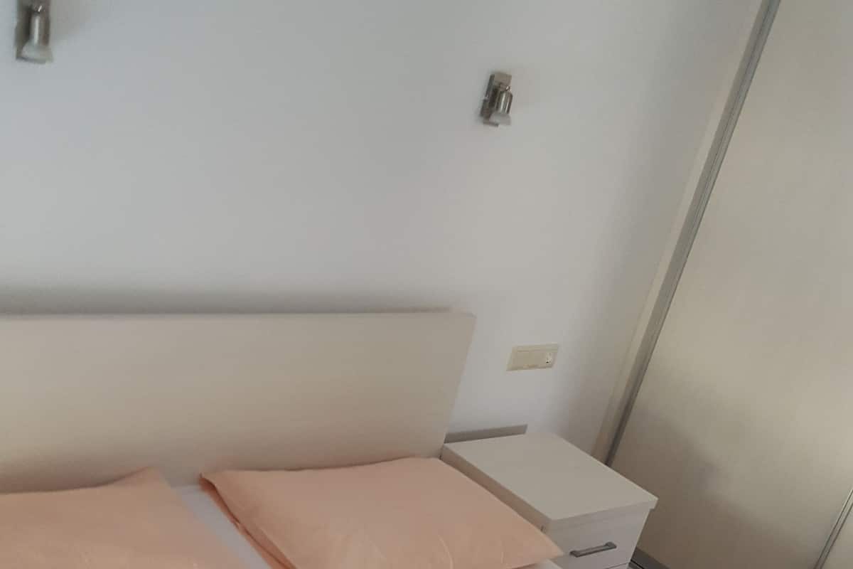 A neatly made bed with white and peach bedding, a wooden floor, and a white nightstand with a drawer.
