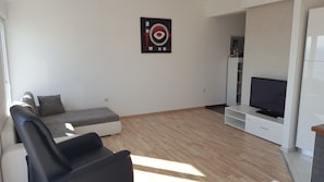 A modern living room with a sofa, a black armchair, a flat-screen TV on a white stand, and a wall-mounted artwork. - Apartman Ivan (Podstrana)