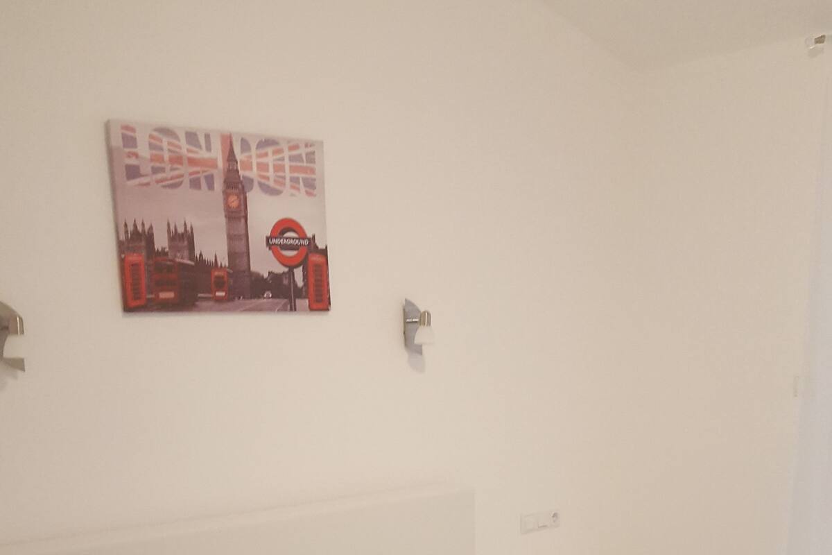 A bedroom with a bed, pillows, a wall-mounted artwork of London landmarks, and a nightstand.