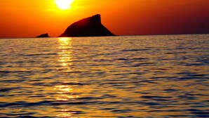Un coucher de soleil sur une mer calme, avec une formation rocheuse imposante se détachant sur le ciel.