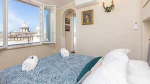 A hotel room with a bed, a window with a view of a dome and buildings, a wall-mounted air conditioner, and a wall-mounted light fixture. - Rental in Rome Ceaser Penthouse (Rome)