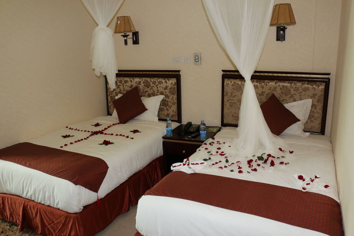 Two double beds with white and brown bedding, a nightstand with a phone and water bottles, and a wall-mounted lamp.