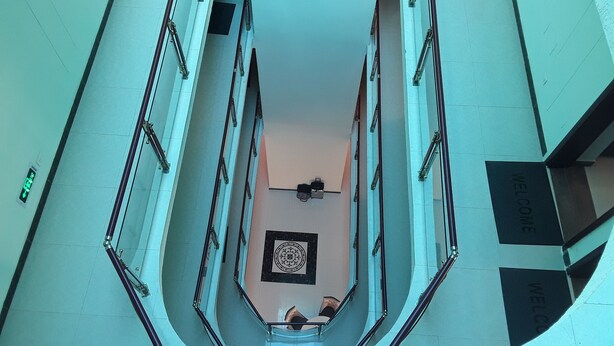 A high-angle view of a modern building's interior, featuring a central open space with a decorative floor pattern, surrounded by glass railings and metal staircases.