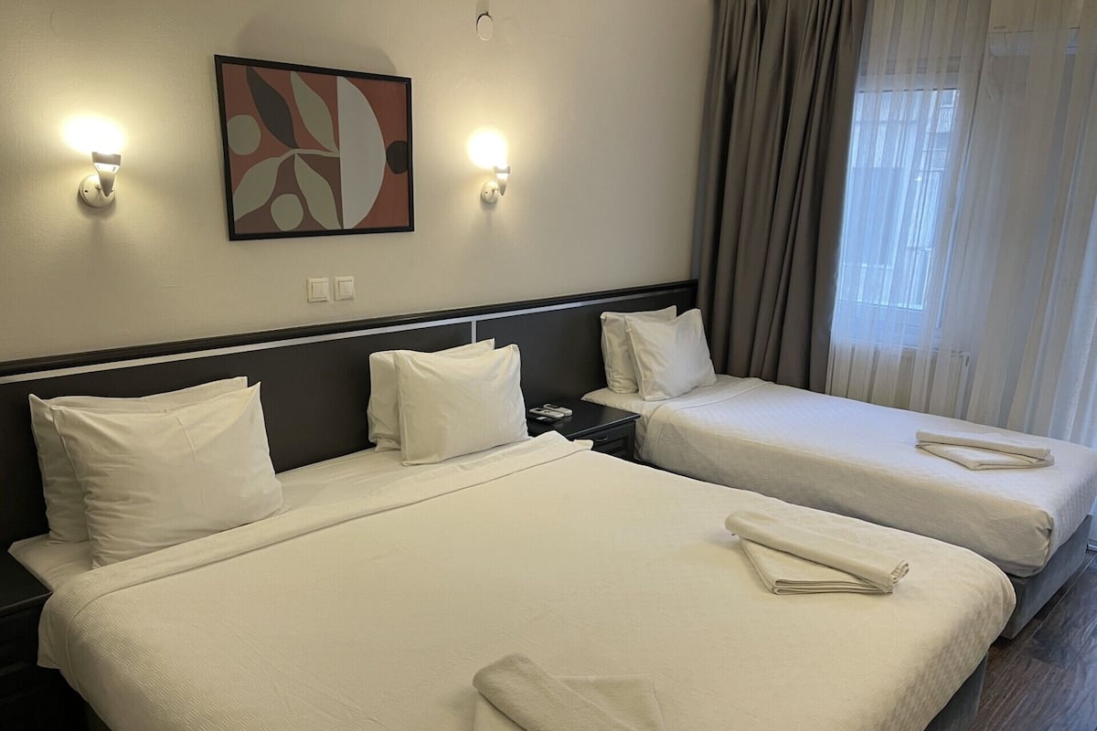A hotel room with two beds, a nightstand, a wall-mounted abstract painting, and a window with curtains.