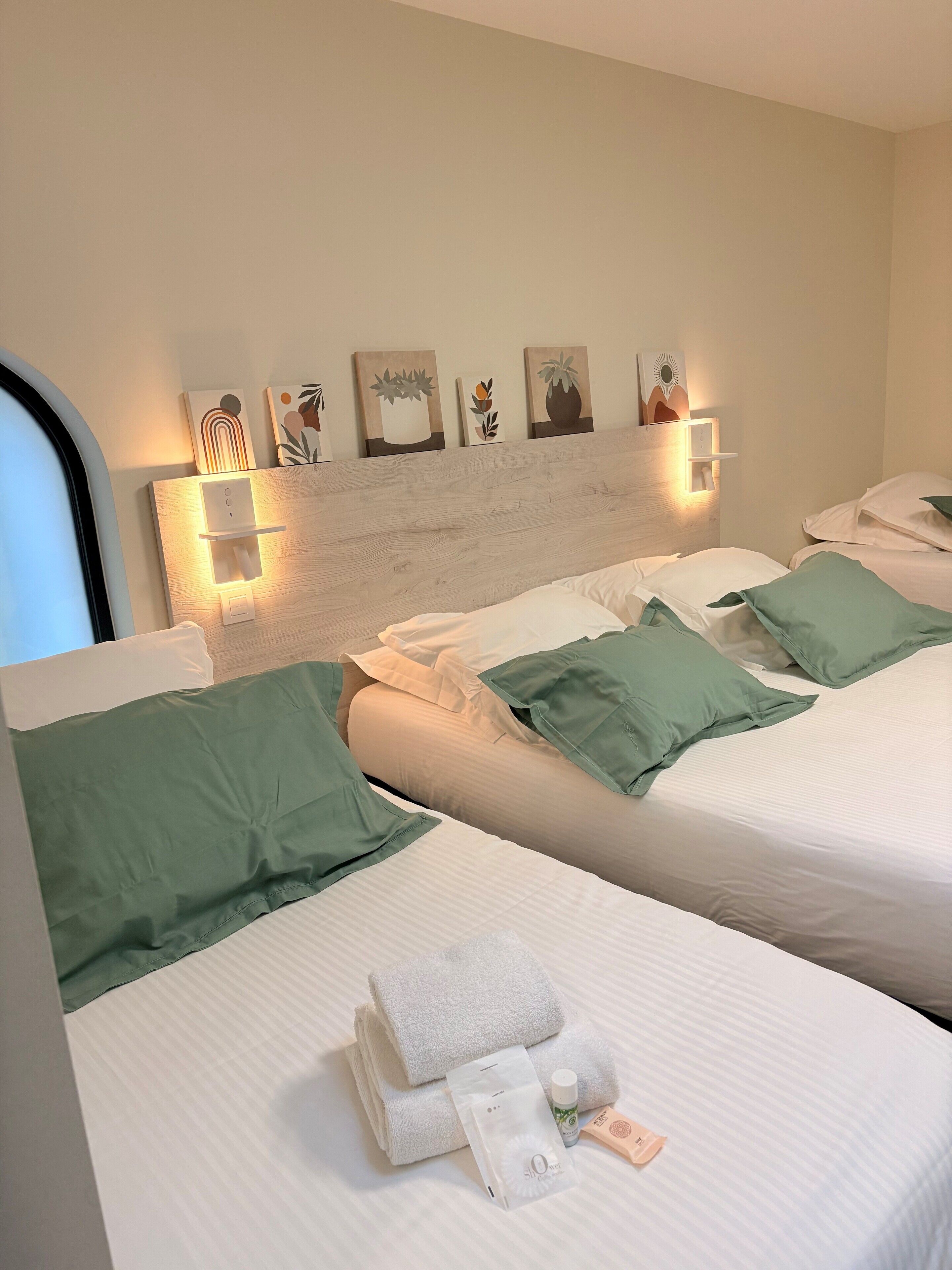 A hotel room with two beds, green pillows, white linens, and a wall-mounted light fixture with artwork.