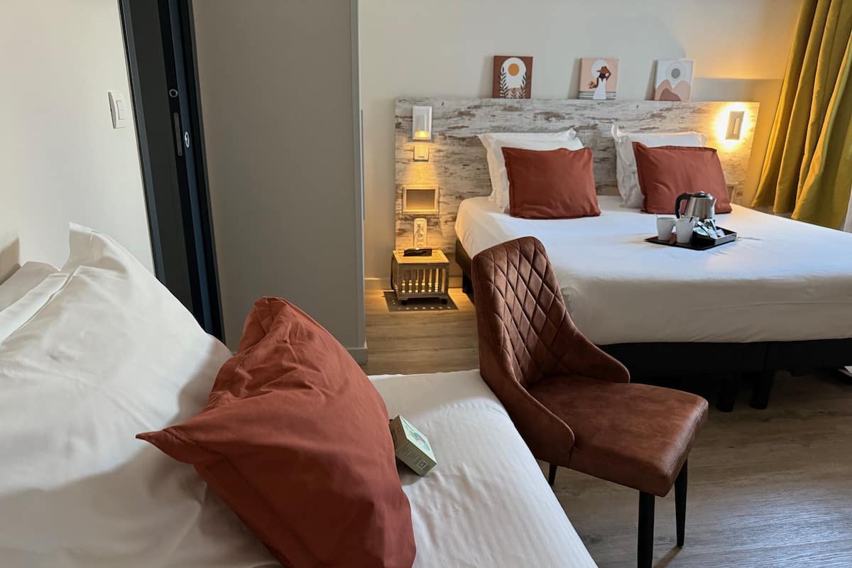 Une chambre d’hôtel avec deux lits, un fauteuil marron et une table de chevet avec une lampe.