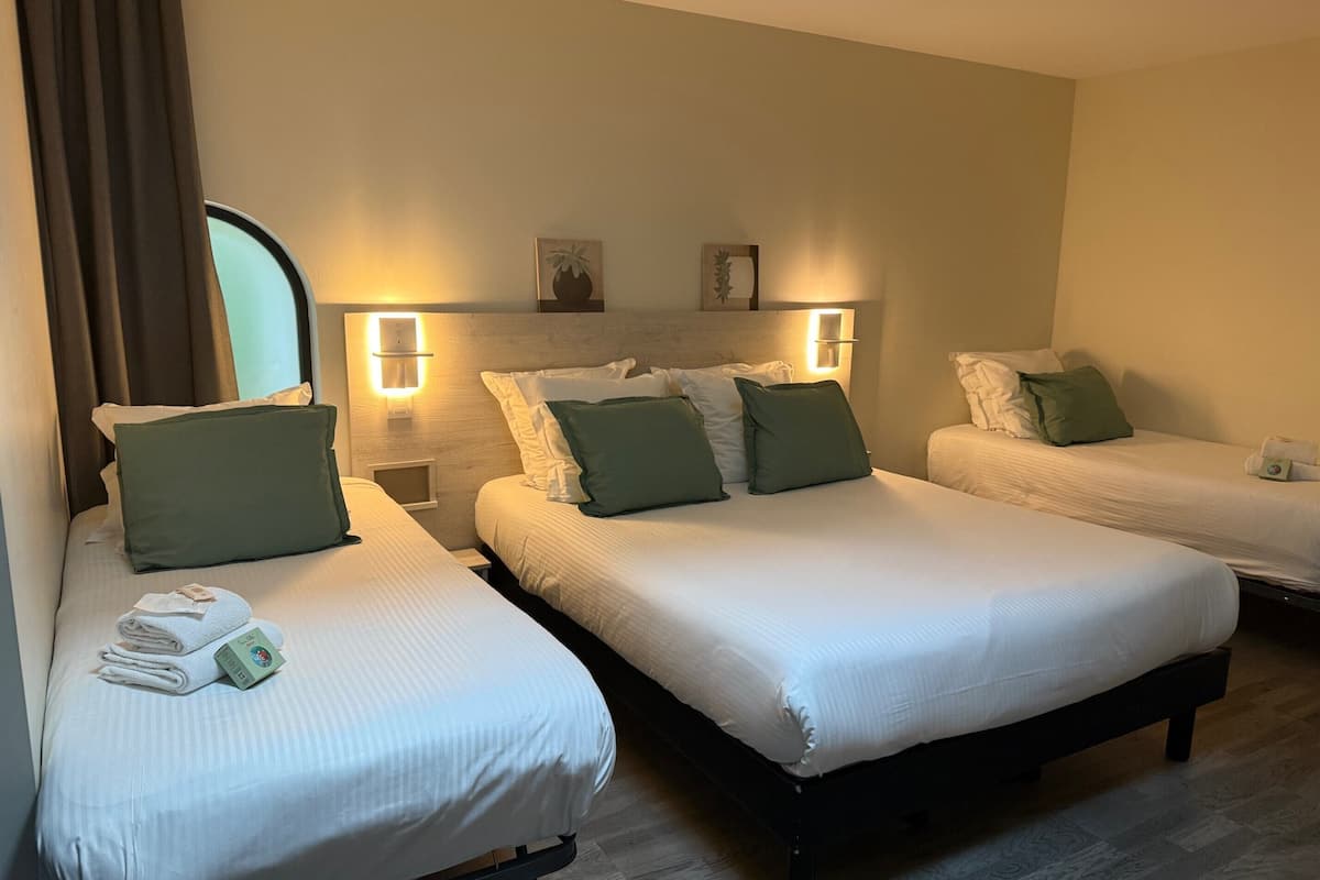 Une chambre d’hôtel avec deux lits simples, chacun agrémenté de draps blancs et d’oreillers verts. Au-dessus de chaque lit, on trouve des appliques murales, et une petite table sur laquelle sont posés une serviette et une carte.