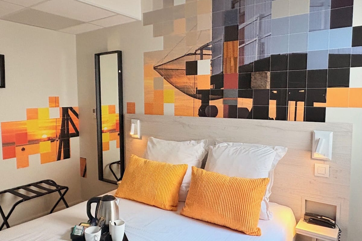 Une chambre d’hôtel avec un lit, deux oreillers, une table de chevet avec un téléphone, un plateau avec des tasses et une cafetière, et un mur orné d’une œuvre d’art géométrique.