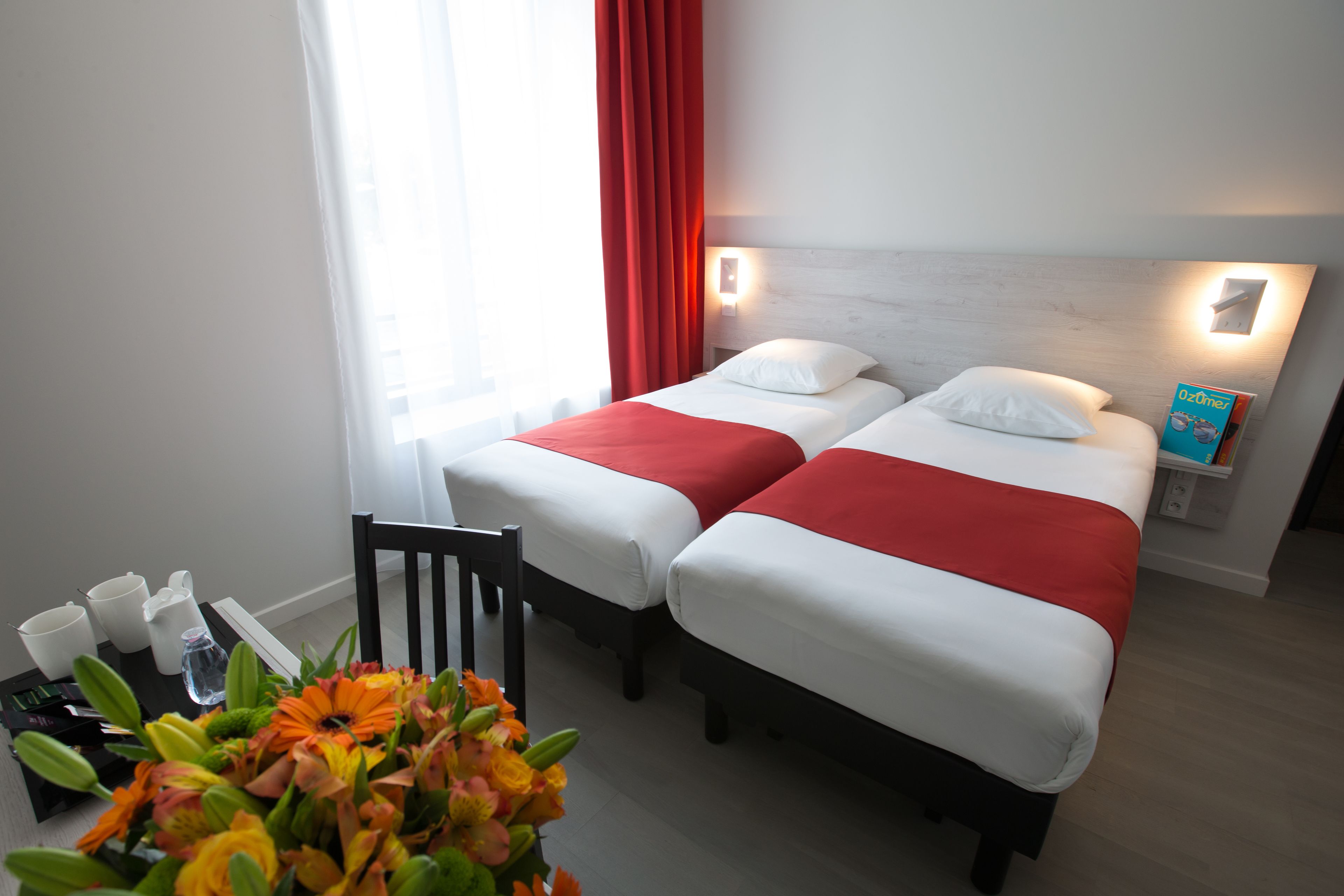 Een hotelkamer met twee bedden, een houten hoofdbord, een nachtkastje met een lamp, een raam met rode gordijnen en een bloemenvaas op de vloer.