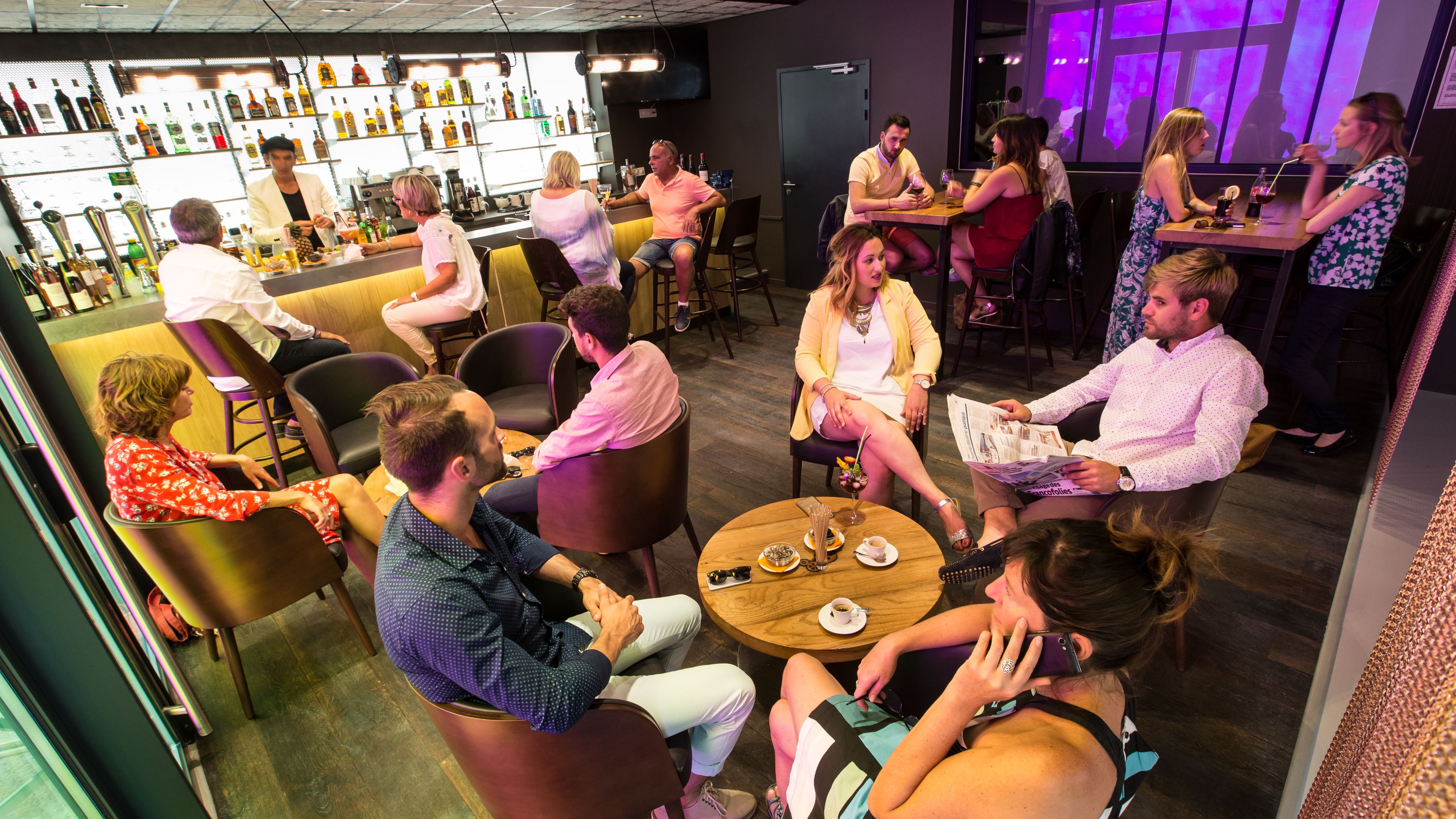 Zona de bar con mesas redondas, sillas y una barra con diferentes botellas.
