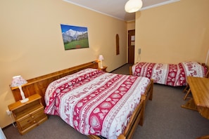 A hotel room with two beds, wooden furniture, a lamp, a picture of mountains, and a door.