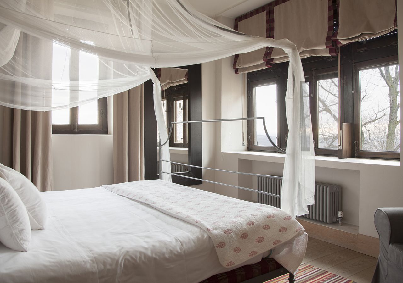 A bedroom with a bed, a window with curtains, a radiator, and a chair.