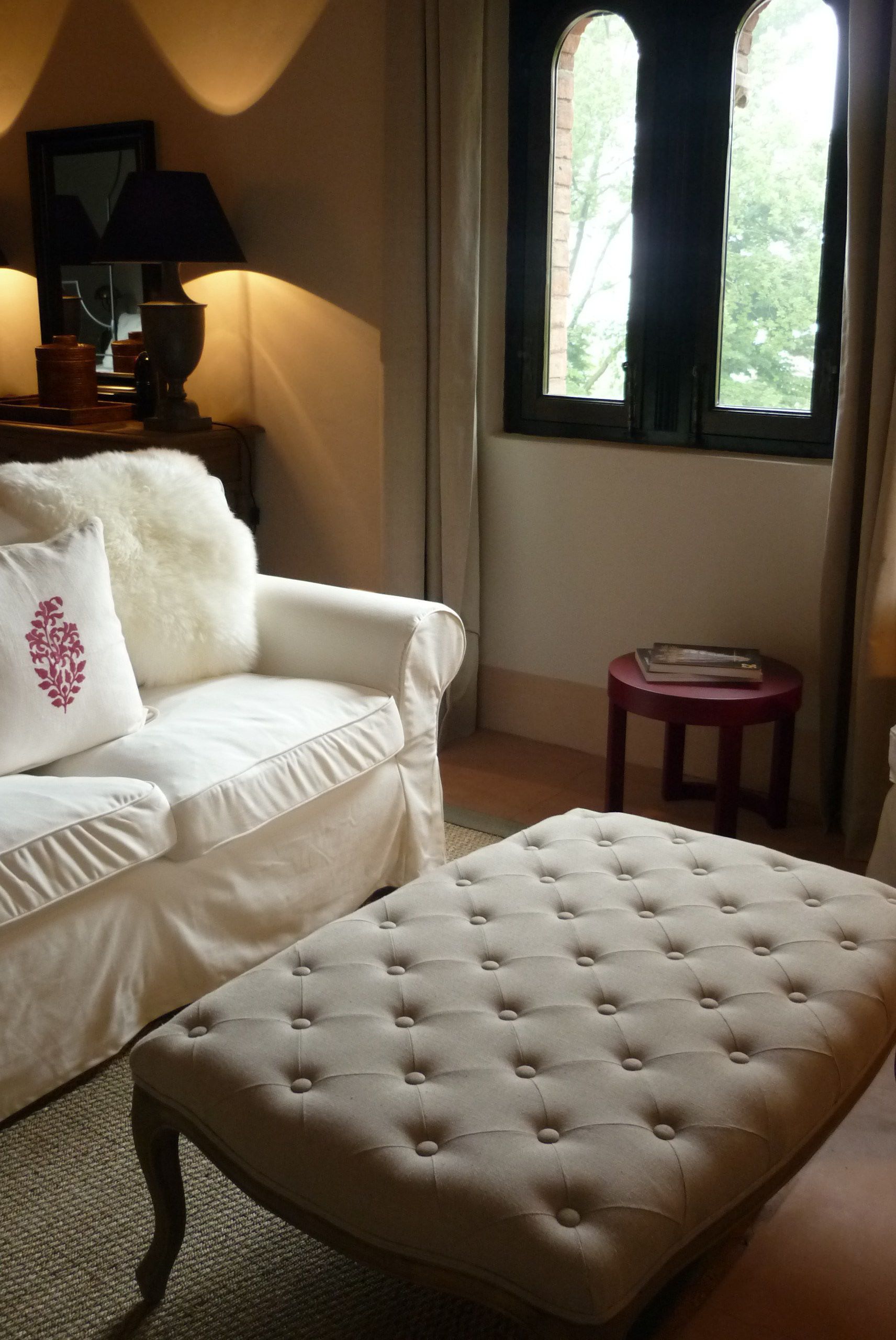 Une pièce avec un canapé blanc, un pouf capitonné, une petite table en bois et une fenêtre donnant sur des arbres.