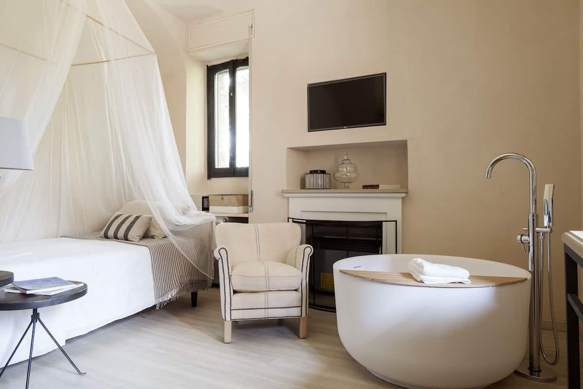 A bedroom with a bed, a chair, a TV, a fireplace, and a bathtub.