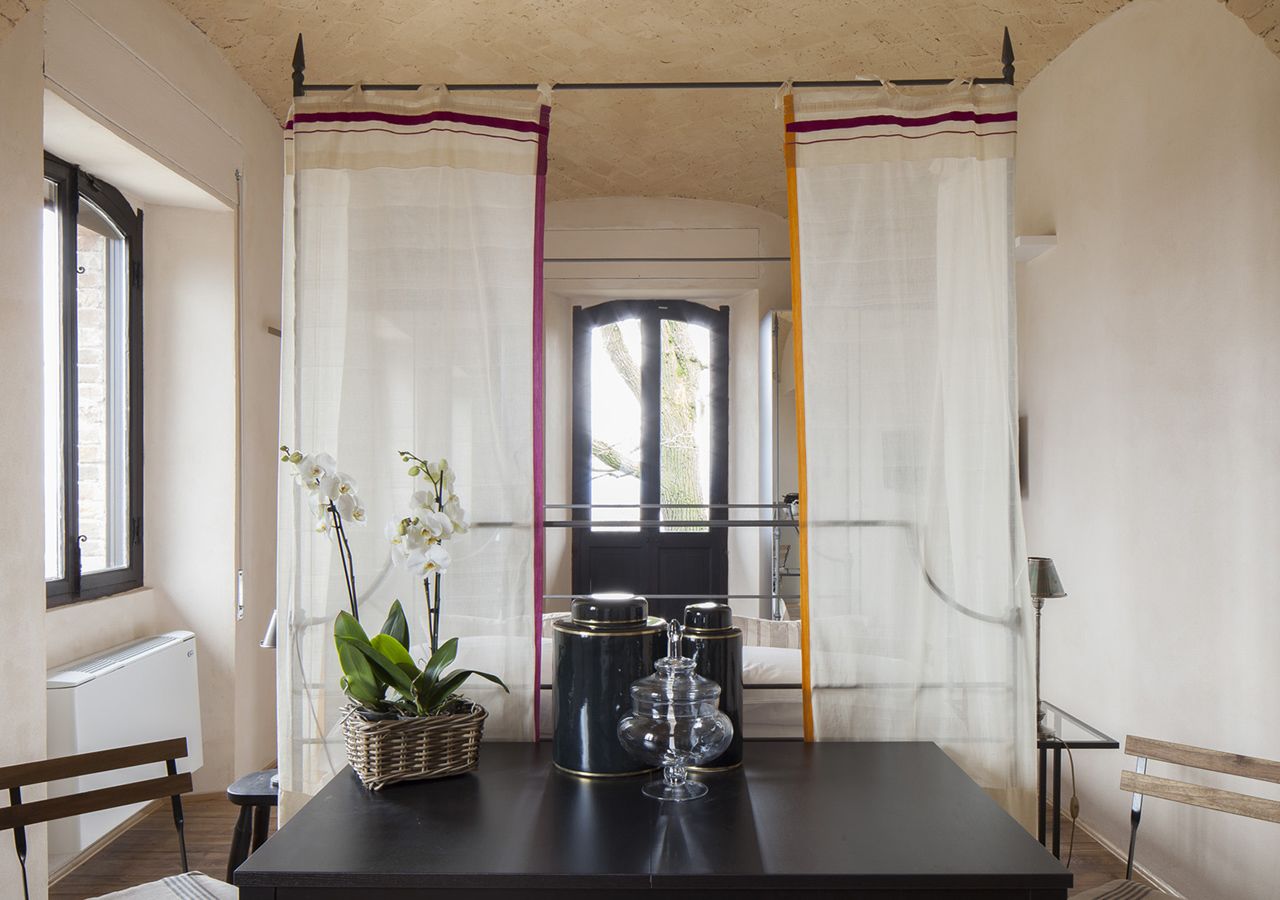 A room with a black table, white curtains, a window, a radiator, a wicker basket with plants, and a lamp.