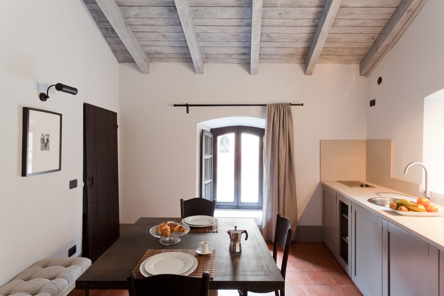 A dining area with a table set for two, a window with curtains, a kitchen with a sink and fruit, and a framed picture on the wall.