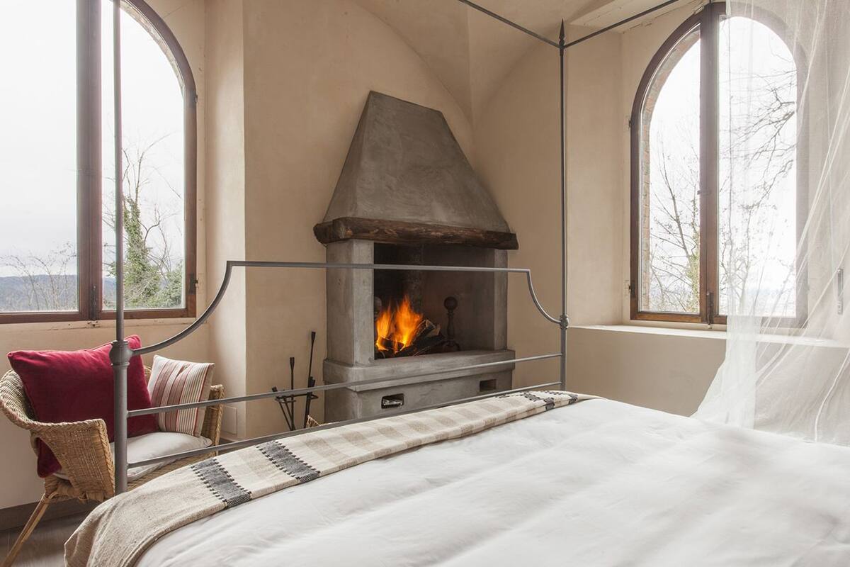 A bedroom with a stone fireplace, a bed, a wicker chair, and a view through large windows.