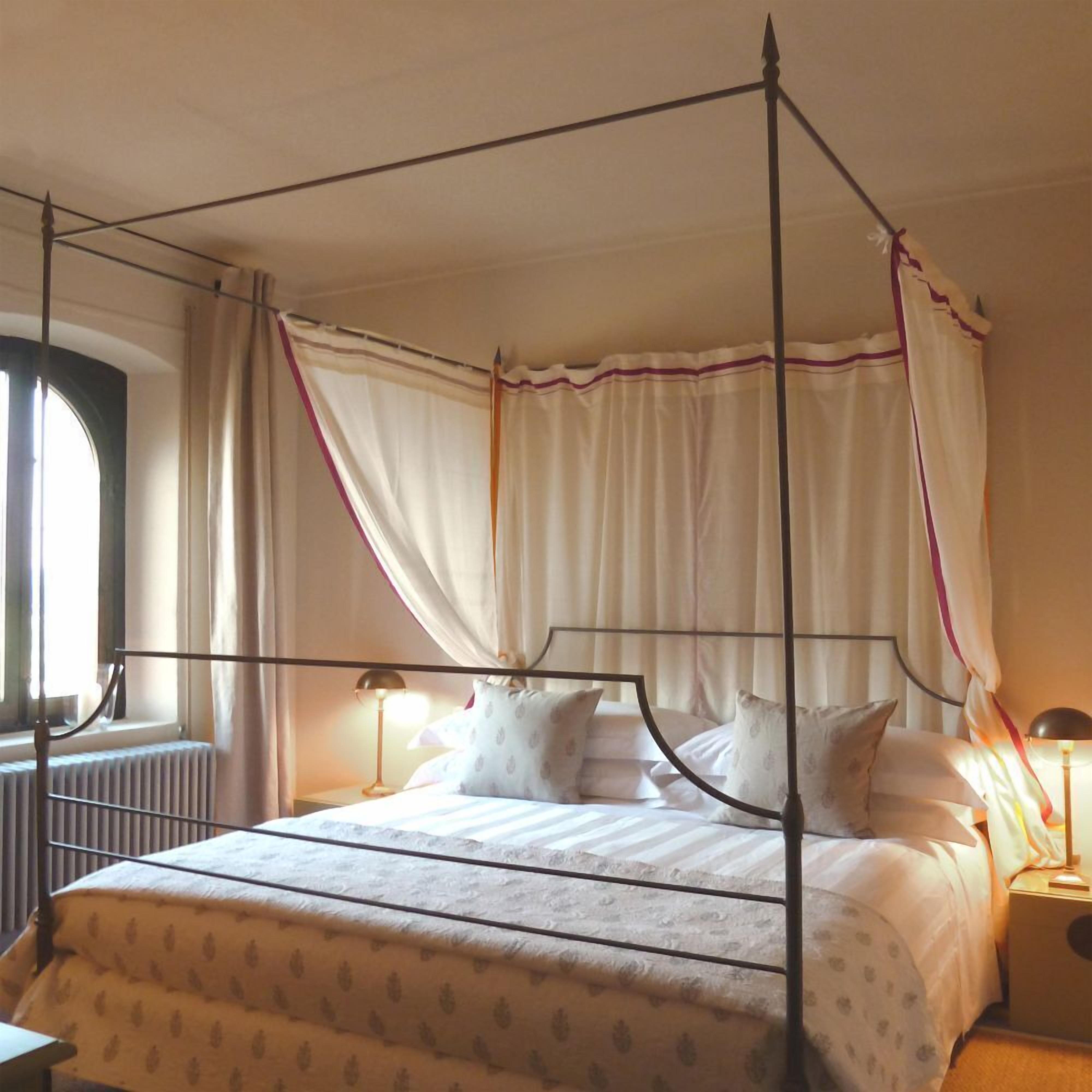 A four-poster canopy bed with white bedding and patterned pillows, a bedside lamp, and a window with curtains.