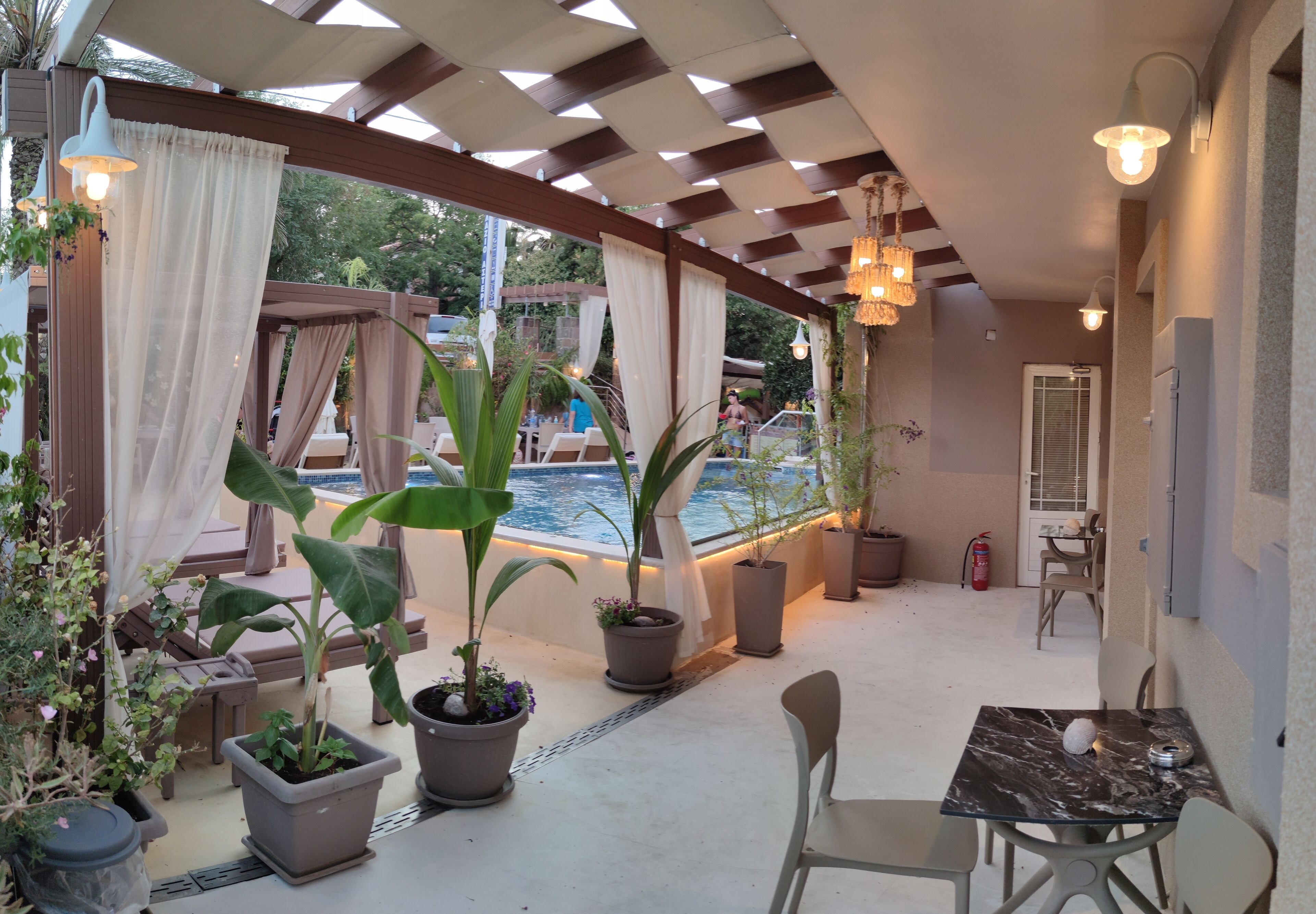A covered outdoor patio with potted plants, a pool, and a table with chairs.