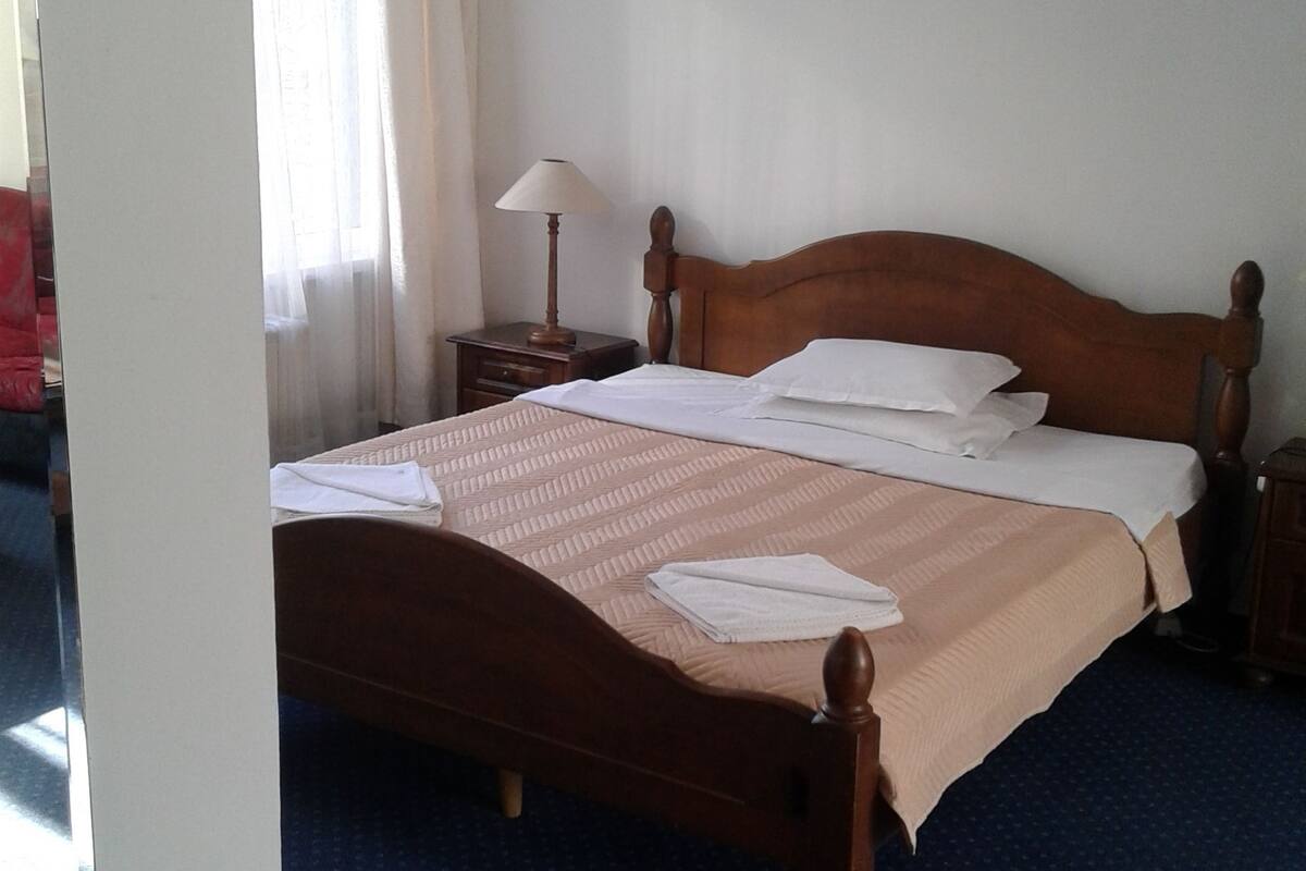 Una cama bien hecha con cabecera de madera, una mesita de noche con lámpara y una ventana con cortinas traslúcidas.