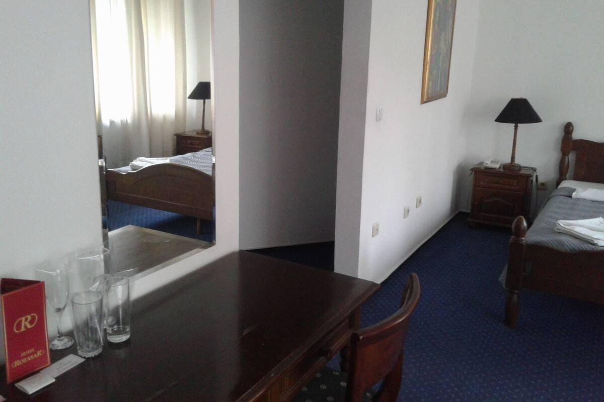 Habitación de hotel con una cama de madera, una mesita de noche de madera con una lámpara, un escritorio con un espejo y una alfombra azul.