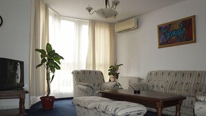 A living room with a sofa, armchair, ottoman, a coffee table, a TV, and two potted plants. - Rotasar Hotel (Sofia)