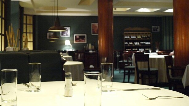 A restaurant interior with a round table set for dining, a hanging pendant light, and a bar area in the background. - Rotasar Hotel (Sofia)