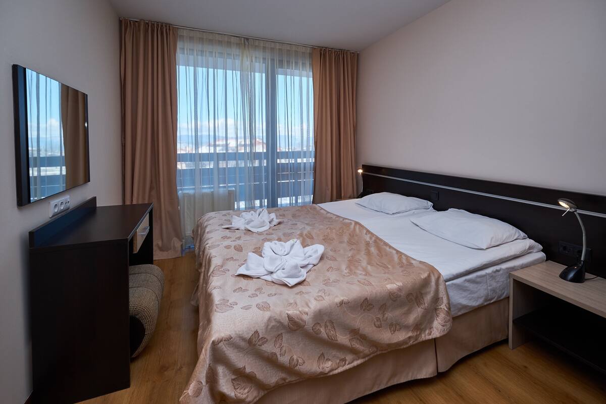 Habitación de hotel con cama, mesita de noche con lámpara, ventana grande con cortinas, televisor de pantalla plana y suelo de madera.