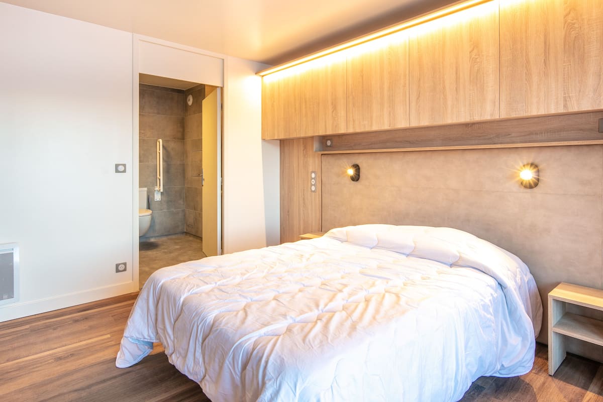 A modern hotel room with a large bed, a walk-in shower, and a wooden headboard with integrated lighting.