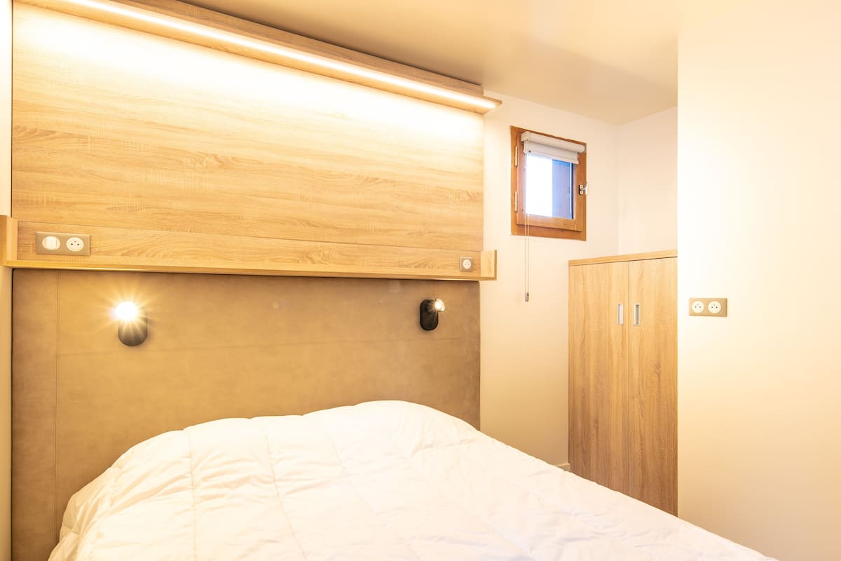 A bedroom with a wooden headboard, a bed with a white duvet, a small window, and a wooden wardrobe.