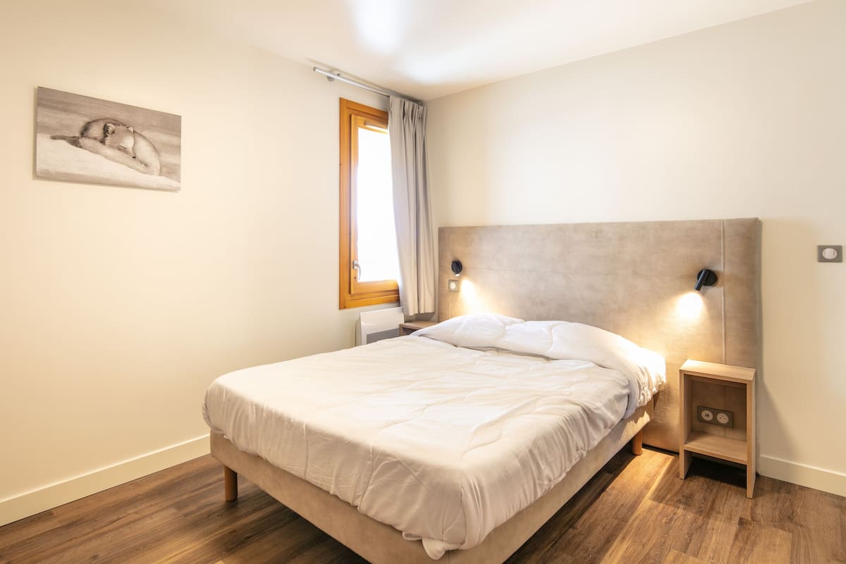 A bedroom with a bed, a nightstand, a wall-mounted lamp, a window with curtains, and a painting on the wall.