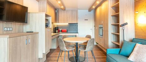 A modern kitchen with wooden cabinets, a dining table with chairs, a built-in oven, and a flat-screen TV mounted above the counter.