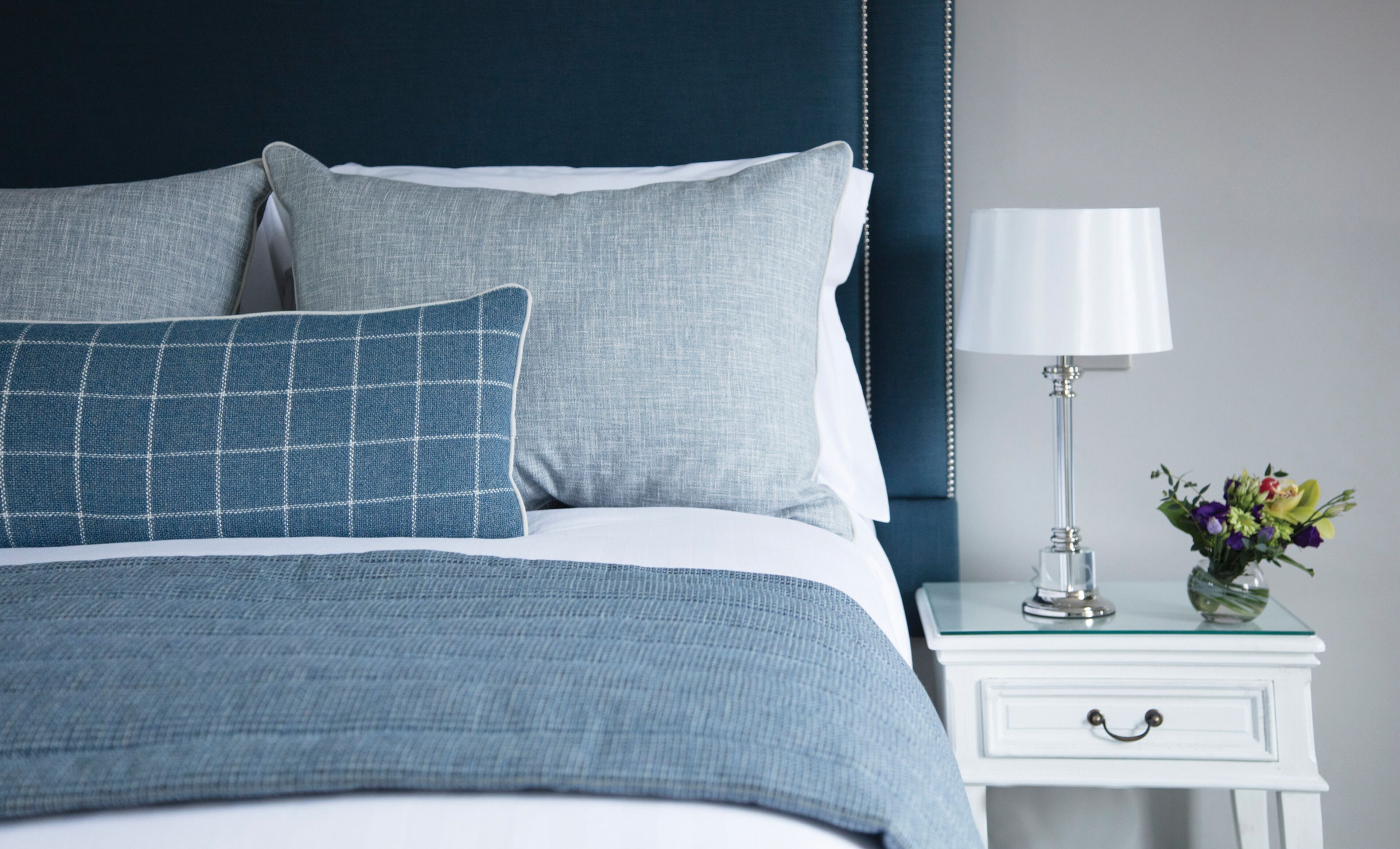 Un letto rifatto con cura, dai colori blu e bianco, un comodino bianco con una lampada e un vaso di fiori, e una testiera blu scuro.