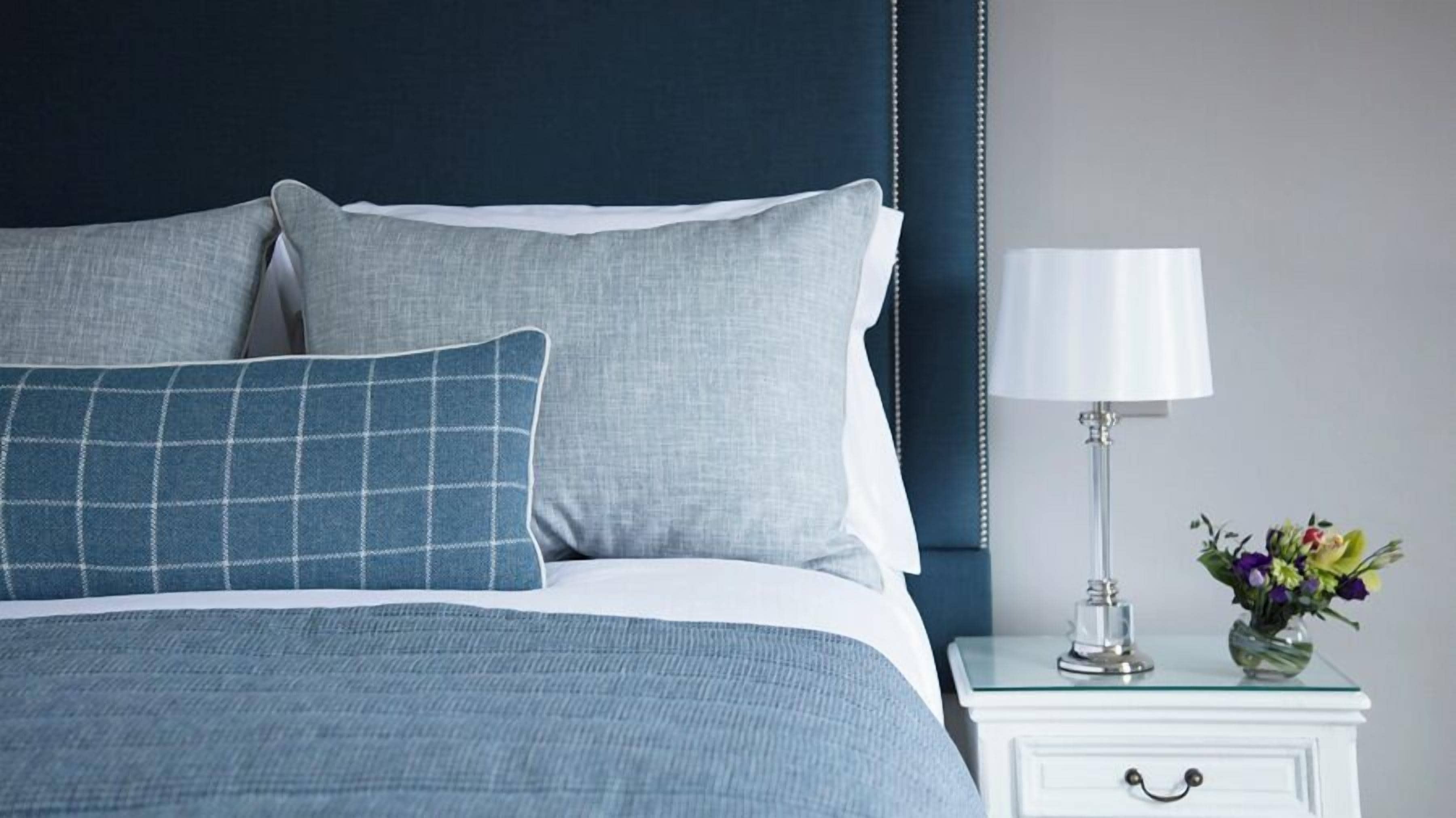 Una cama bien tendida con ropa de cama azul y blanca, una almohada a cuadros azules y blancos y una mesita de noche blanca con una lámpara y un florero con flores.