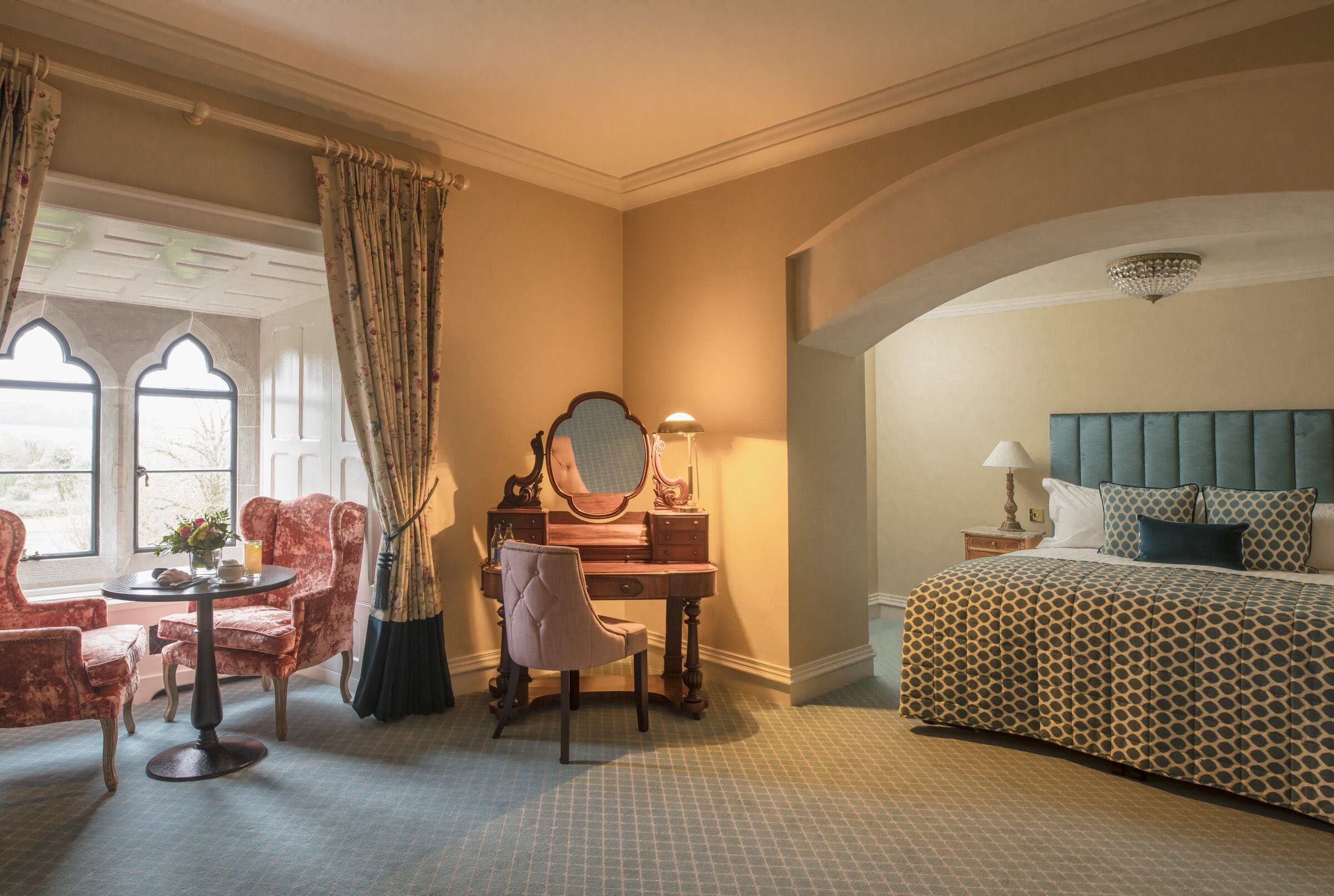 A hotel room with a bed, a desk with a mirror, two chairs, a small table, and a window with curtains.