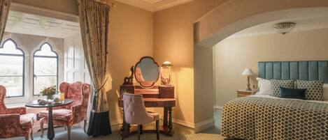 A hotel room with a bed, a desk with a mirror, two chairs, a small table, and a window with curtains.