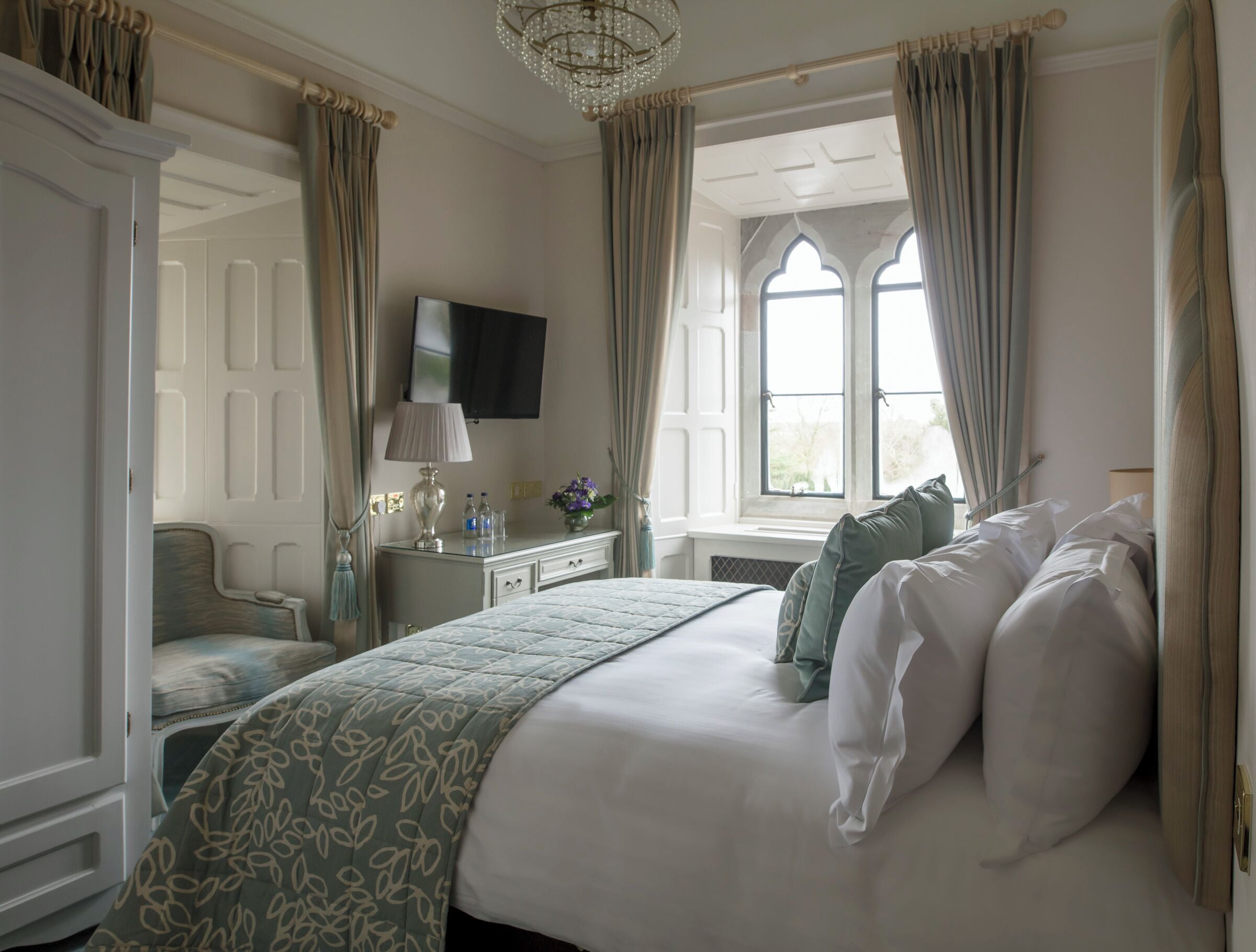 Een hotelkamer met een groot bed, een bureau met een lamp, een stoel, een televisie en een raam met gordijnen.