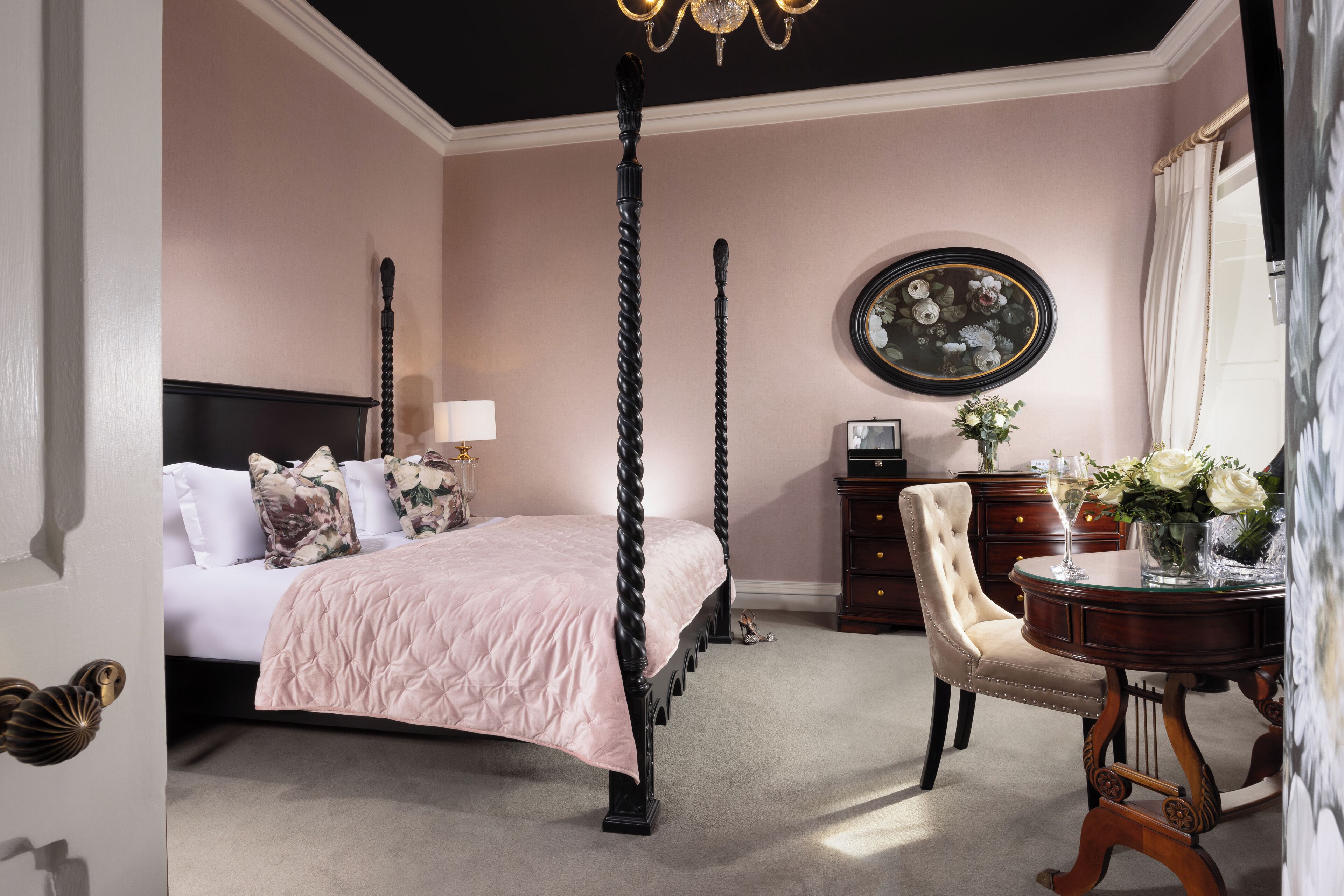 Una cama con dosel con una colcha rosa, una mesita de noche de madera con un jarrón de flores y una mesa redonda con un jarrón de flores.
