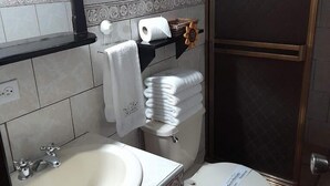 A bathroom with a white sink, toilet, and shower. There is a towel rack with towels and a toilet paper roll.