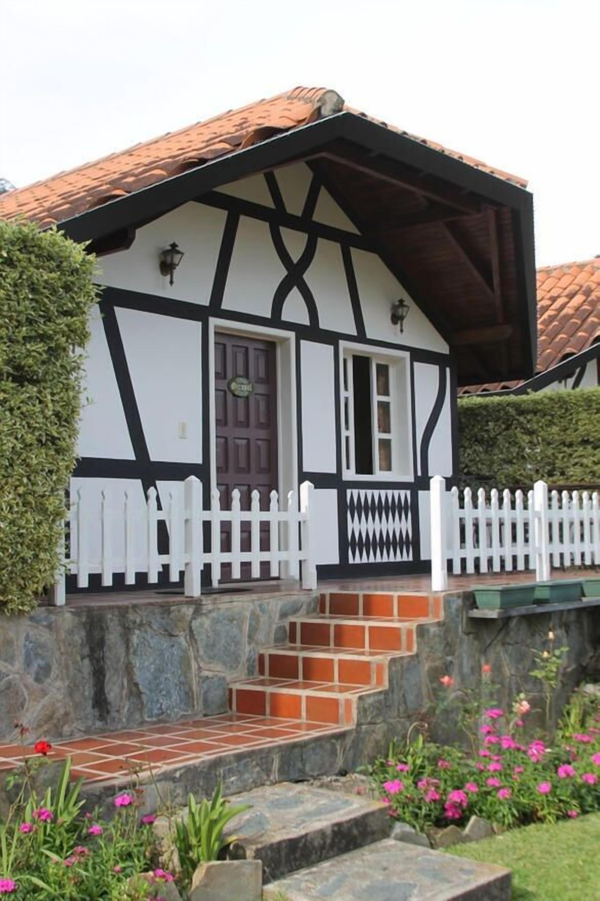 A traditional half-timbered house with a white picket fence, stone steps, and a small garden with pink flowers.