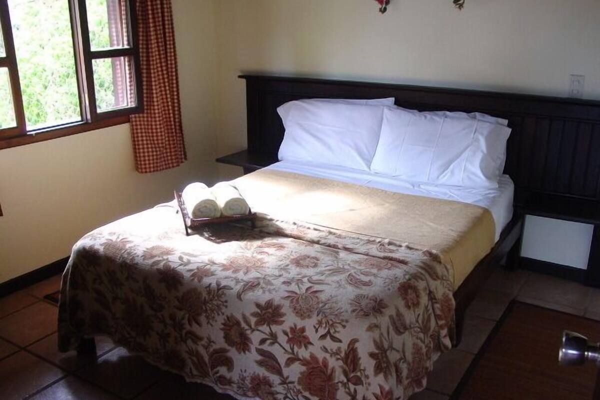 Un lit bien fait, recouvert d’une couvre-lit à motifs floraux, une pile de serviettes enroulées et une fenêtre avec des rideaux.