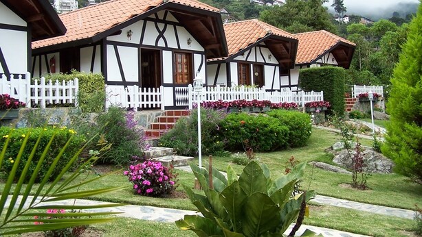 A traditional half-timbered house with a white picket fence, surrounded by well-maintained gardens and a hilly backdrop.