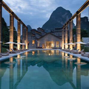 Ein Poolbereich mit Liegestühlen und Sonnenschirmen, ein Gebäude mit großen Fenstern und ein Berg im Hintergrund.